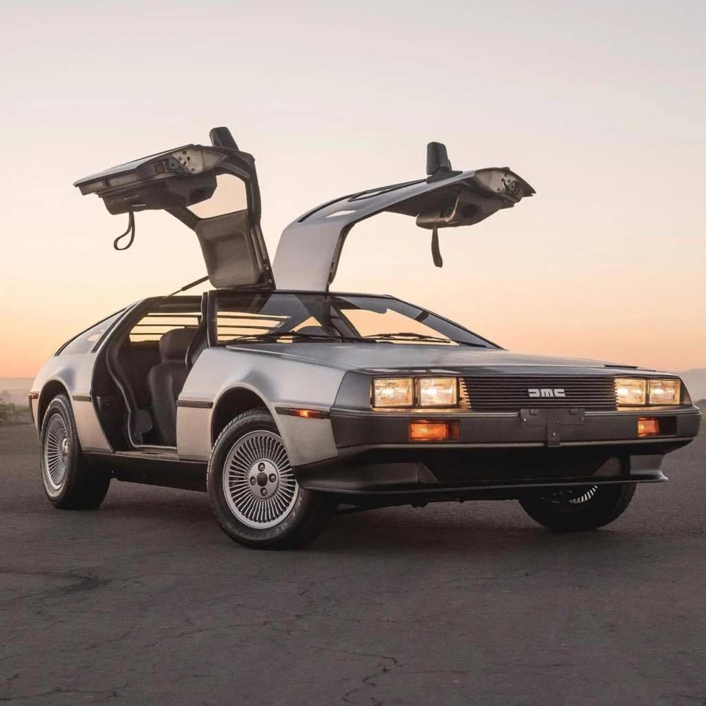 Silver DeLorean car with iconic gull-wing doors open, parked on pavement at sunset, evoking 80s nostalgia and futuristic design.
