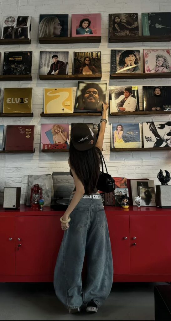 e0da0e49-13b8-4a04-8fd4-428db283961a A person in casual attire reaches for a record on a wall display, surrounded by various album covers. | Sky Rye Design A person in casual attire reaches for a record on a wall display, surrounded by various album covers.