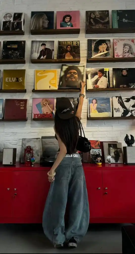 e0da0e49-13b8-4a04-8fd4-428db283961a A person in casual attire reaches for a record on a wall display, surrounded by various album covers. | Sky Rye Design A person in casual attire reaches for a record on a wall display, surrounded by various album covers.