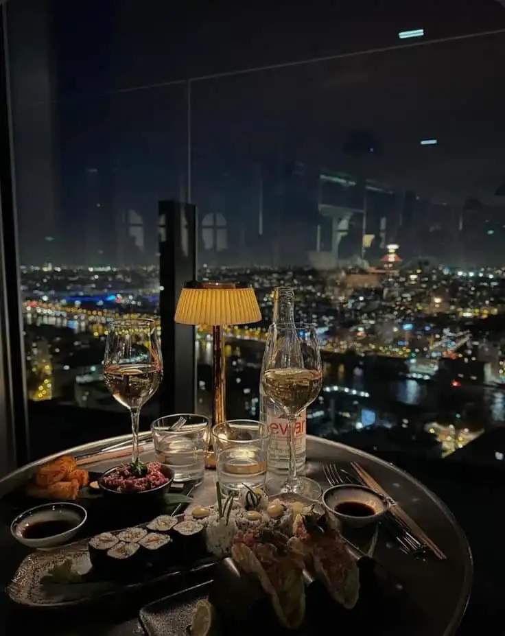 Elegant dining with sushi and wine overlooking a stunning cityscape at night, enhanced by ambient table lighting.