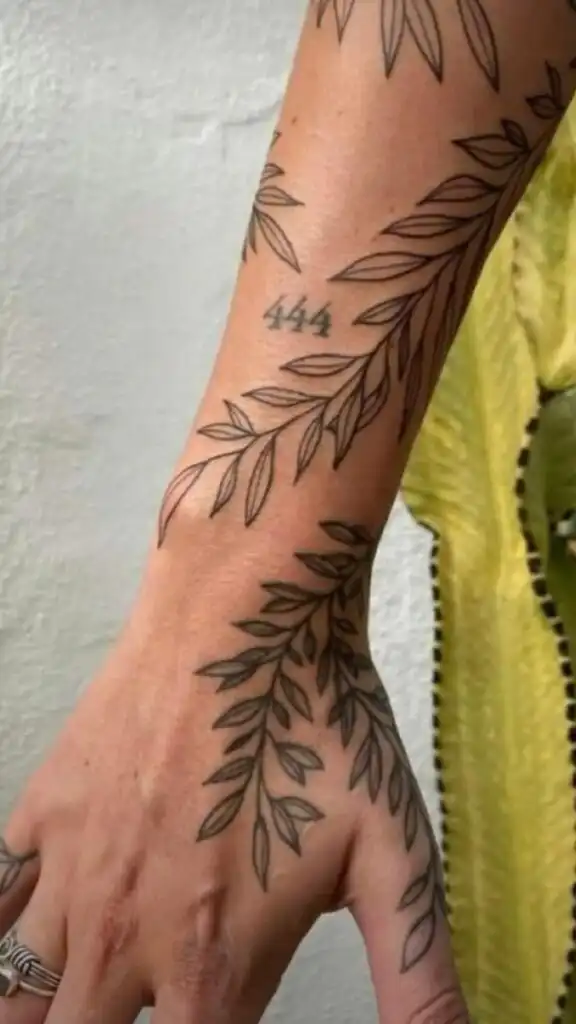 Leafy vine tattoo with 444 on forearm and hand, beside a yellow garment. Minimalist linework design, symbolizing nature and harmony.