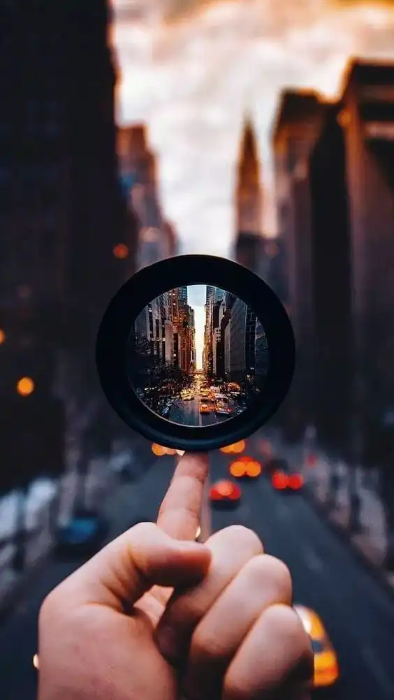 fa364992-7dda-4f61-9950-22ed38b3a2bd City street view focused through a lens held by a hand, creating a sharp contrast between the lens and blurred urban background. | Sky Rye Design City street view focused through a lens held by a hand, creating a sharp contrast between the lens and blurred urban background.