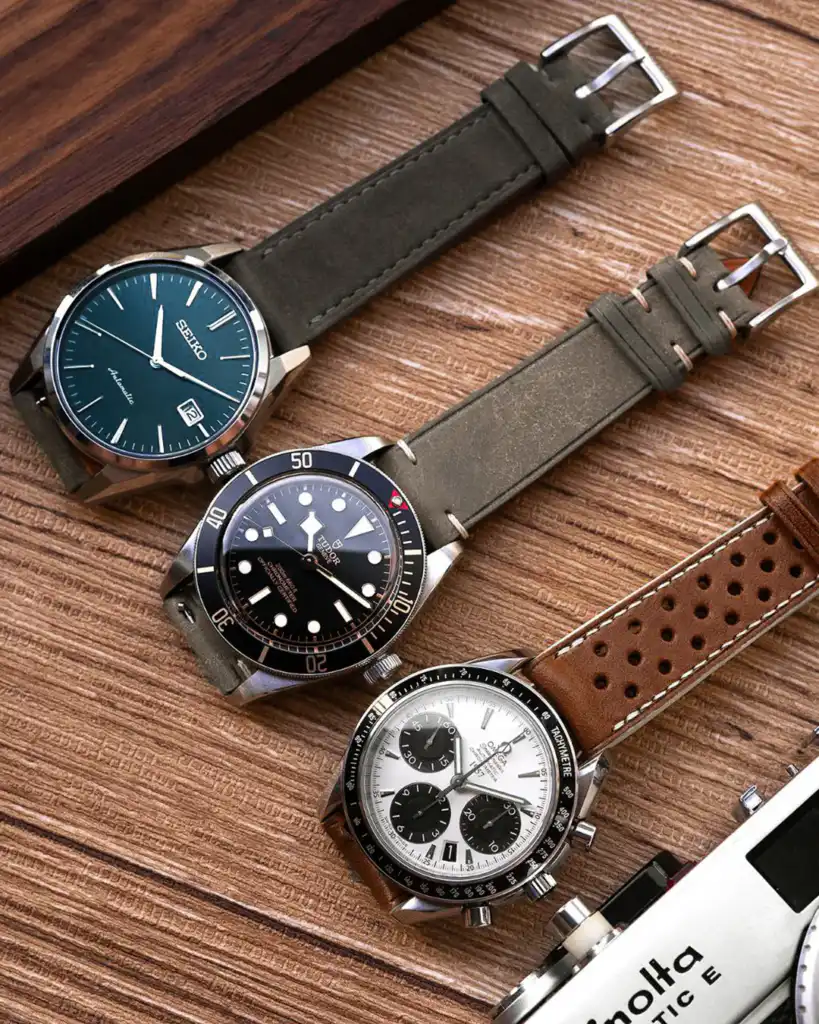 Three stylish watches with leather straps arranged on a wooden surface: a classic, a diver, and a chronograph model.