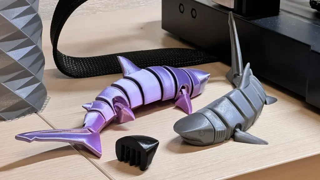 3D printed articulated sharks in purple and gray displayed on a wooden table next to a 3D printer.