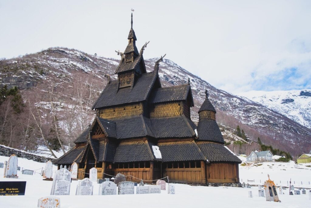 Historic wooden stave church in snowy landscape with mountains, highlighting Norwegian architecture and cultural heritage.
