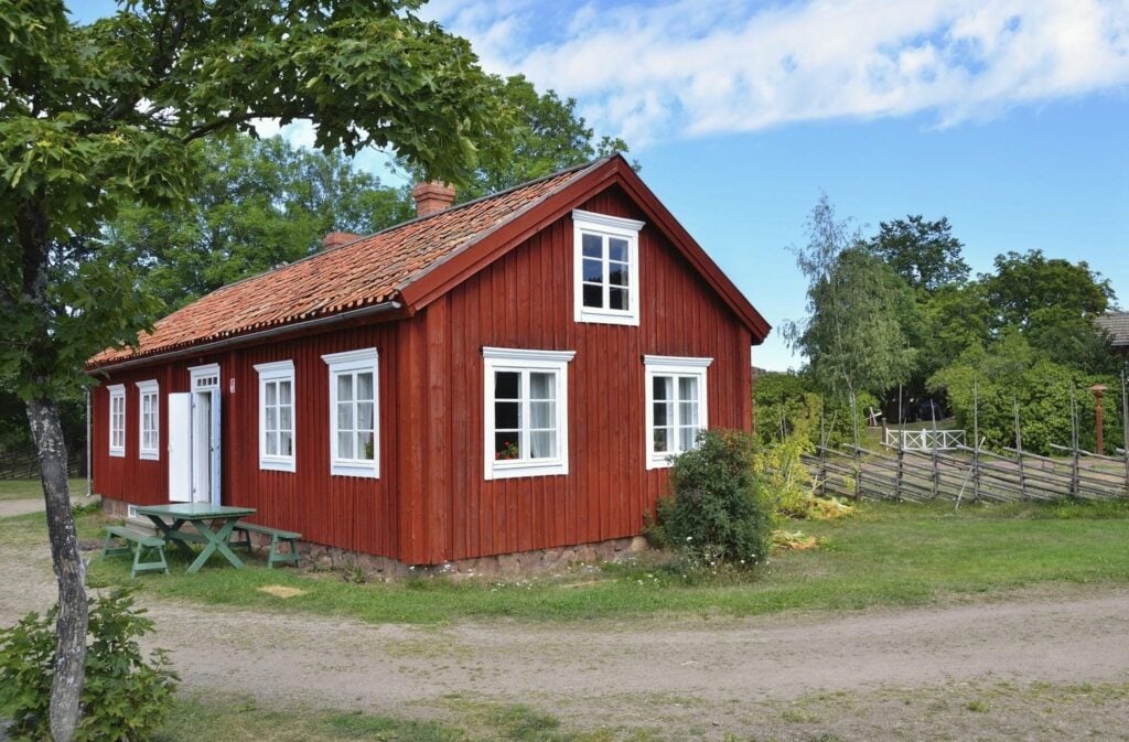 Red wooden cottage with white windows surrounded by green trees, grass, and a clear blue sky, showcasing rustic charm.