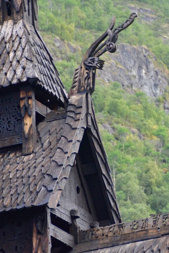 Intricate dragon carvings on the rooftop of a historic wooden stave church in Norway, surrounded by lush greenery and rocky terrain.