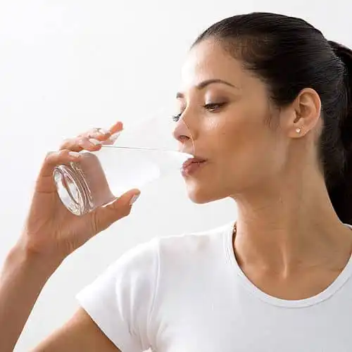 Woman drinking a glass of water, promoting hydration and healthy lifestyle, wearing a white shirt in a bright setting.