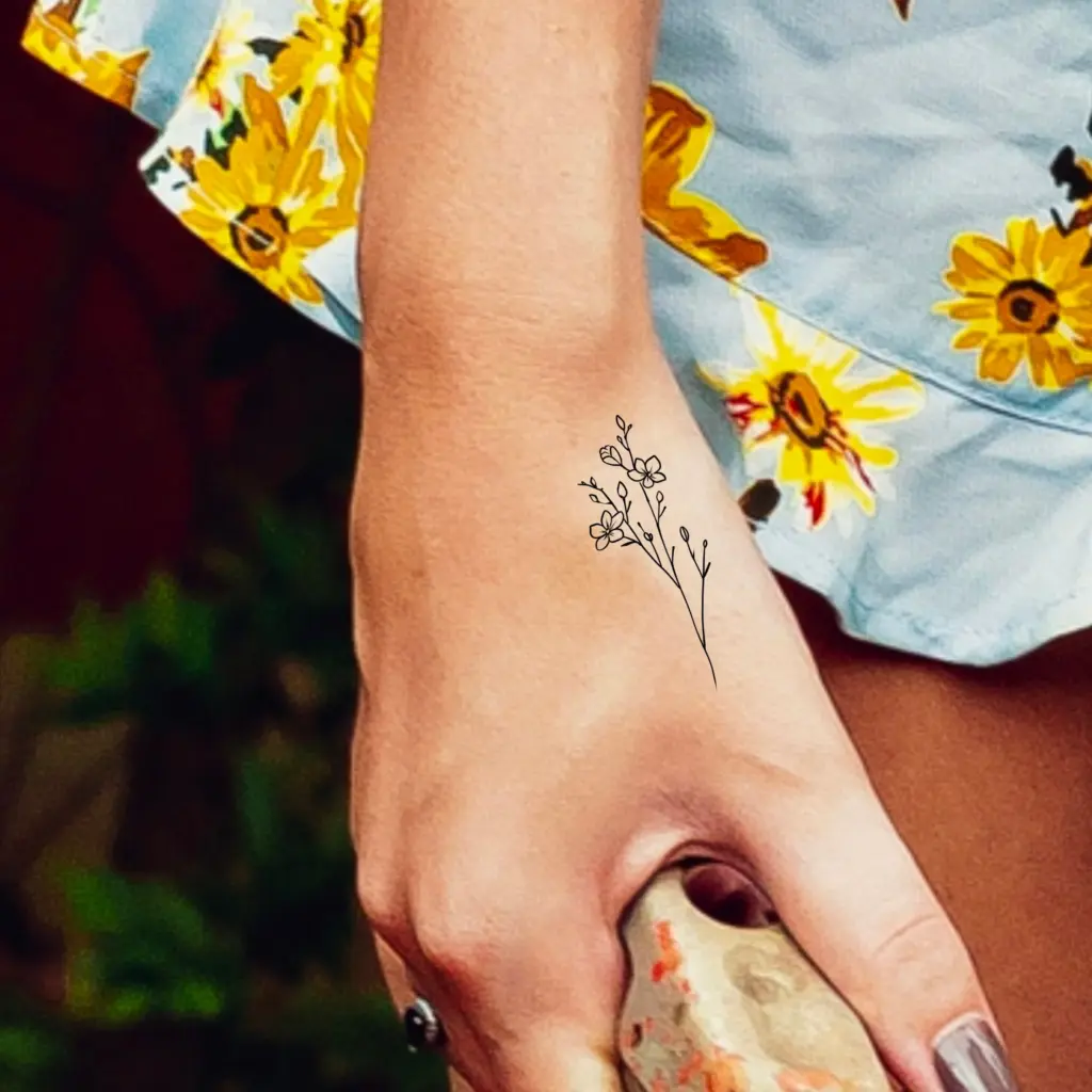 Delicate floral wrist tattoo on a hand holding a rock, paired with a dress featuring vibrant yellow flowers.