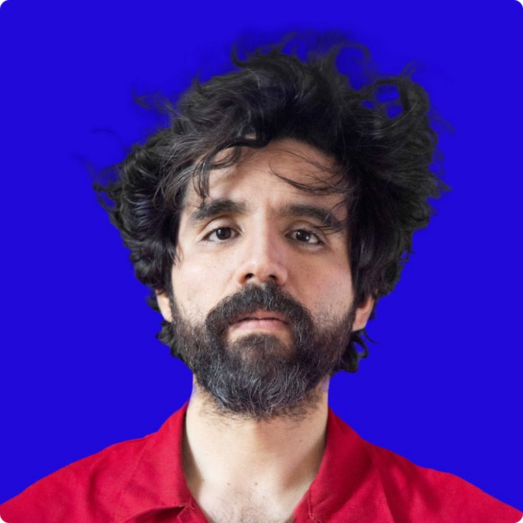 Bearded man in a red shirt with tousled hair against a bright blue background, looking directly at the camera.
