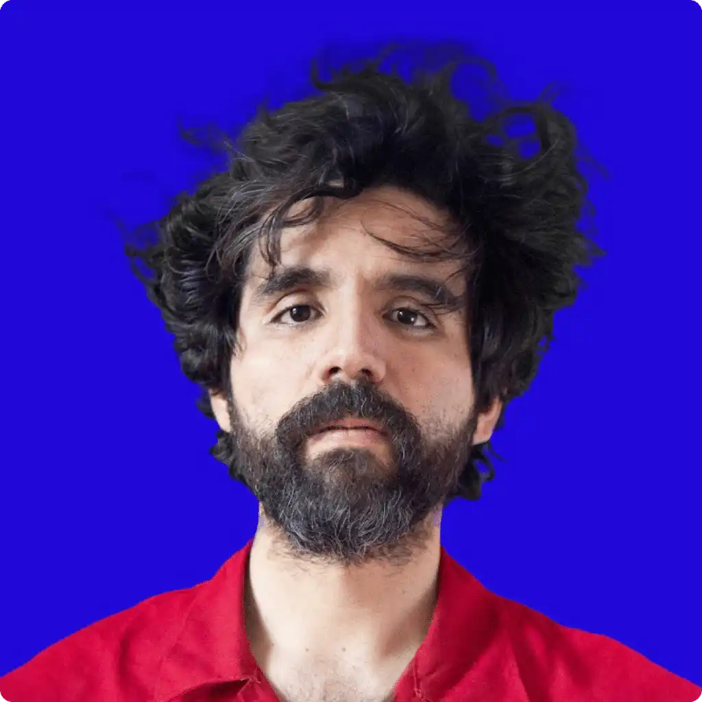 Bearded man in a red shirt with tousled hair against a bright blue background, looking directly at the camera.