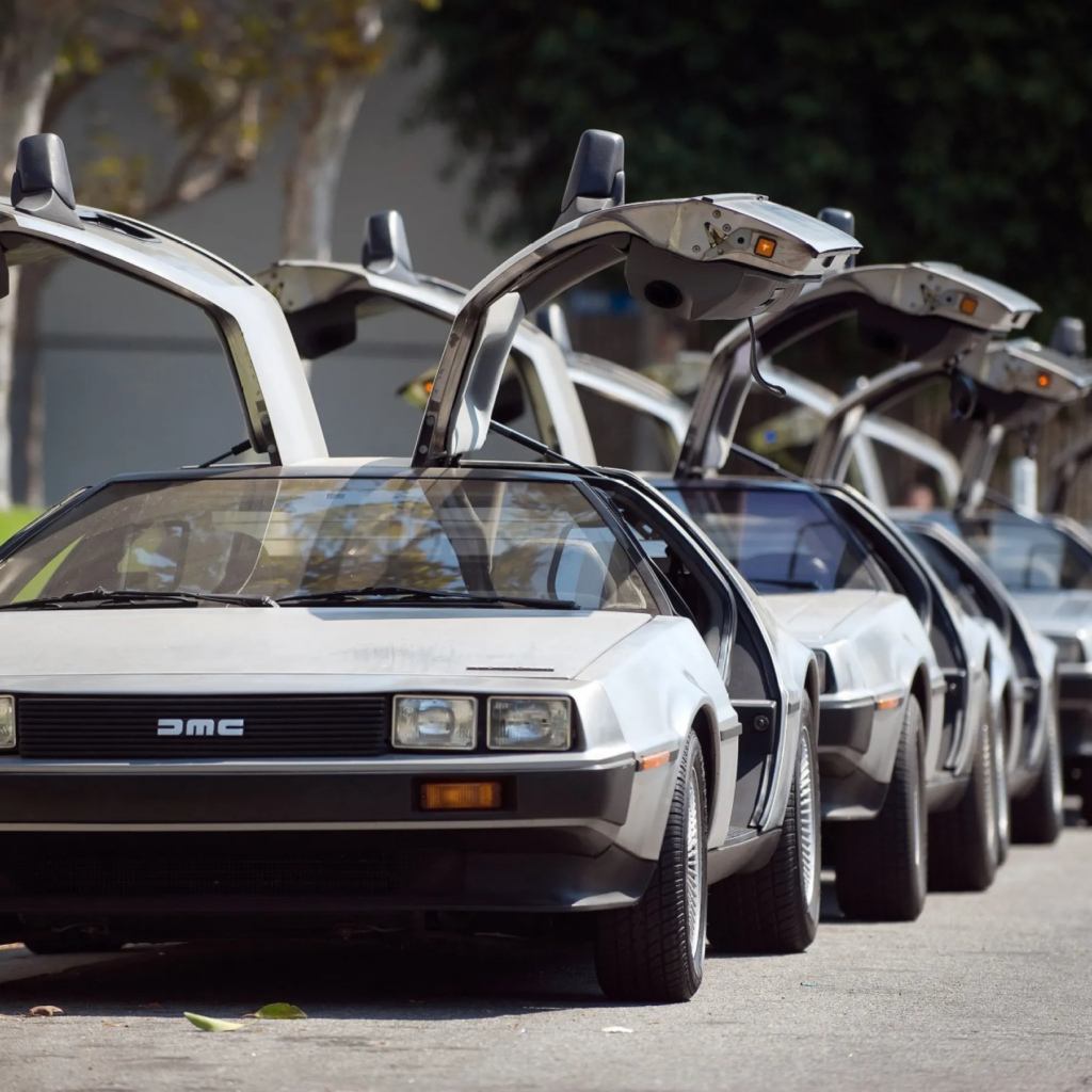Row of classic DeLorean cars with iconic gullwing doors open, parked on a street, capturing retro automotive style.