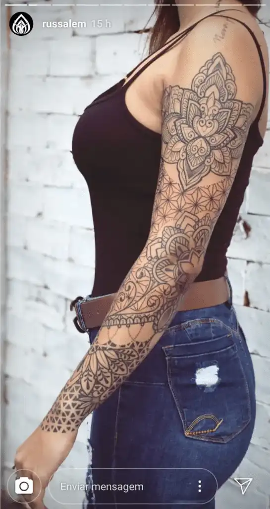 Arm covered in intricate mandala tattoos, woman in black top and jeans against a white brick wall background.