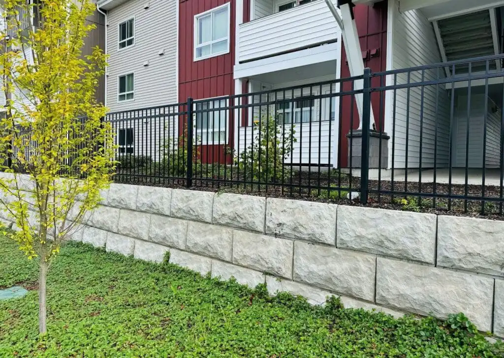 image Modern apartment building with a black metal fence, stone retaining wall, and vibrant green landscaping. Urban residential architecture. | Sky Rye Design Modern apartment building with a black metal fence, stone retaining wall, and vibrant green landscaping. Urban residential architecture.
