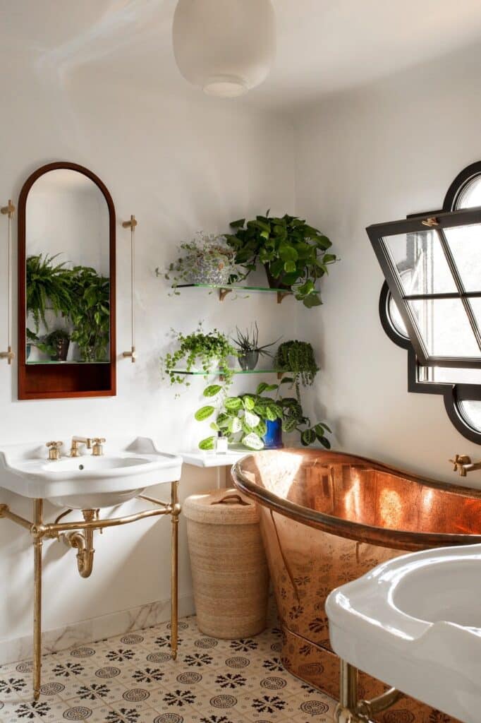 Elegant bathroom with copper bathtub, white sink, gold fixtures, wall mirror, and lush green plants on shelves by a window.