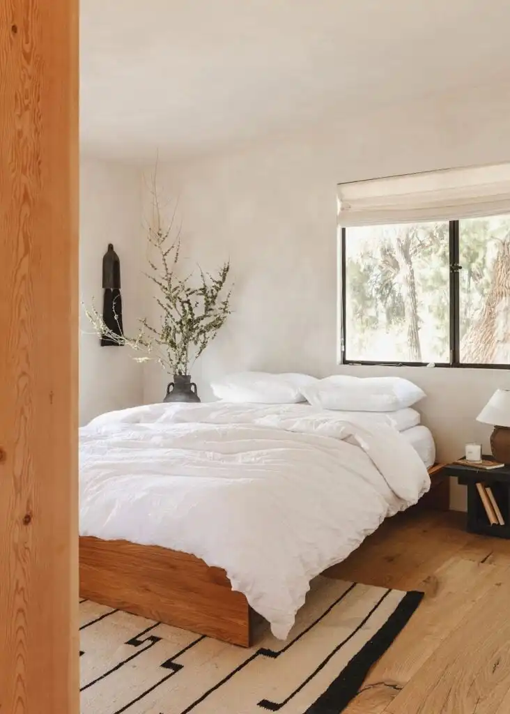 Cozy bedroom with white bedding, wooden floor, large window, and natural decor creating a serene, minimalist atmosphere.