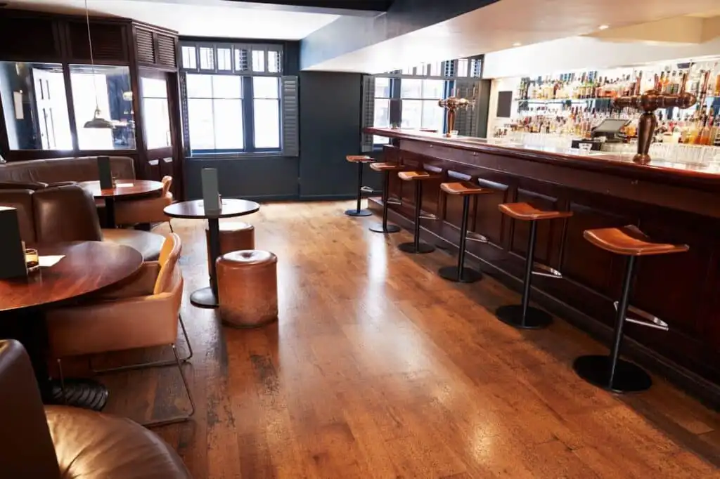 Cozy urban bar interior with wooden furniture, high stools, and tables, featuring a well-stocked bar counter and large windows for natural light.