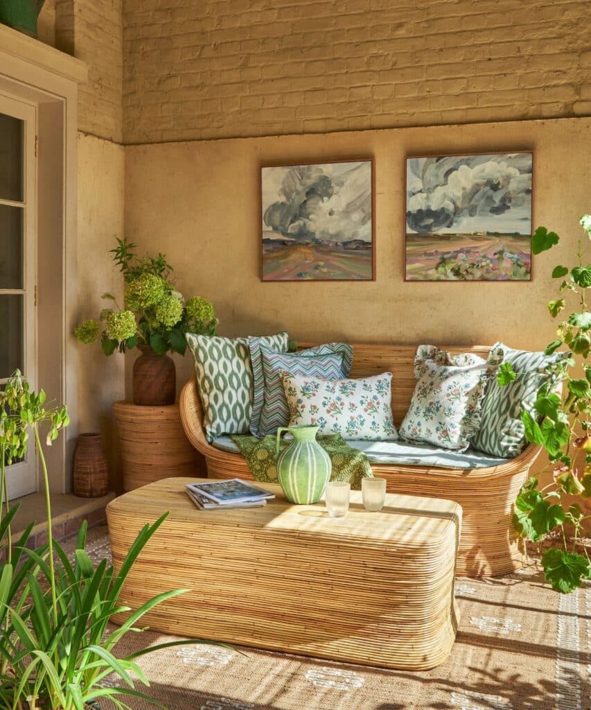 Cozy boho living room with wicker furniture, vibrant cushions, green plants, and landscape paintings on brick wall.