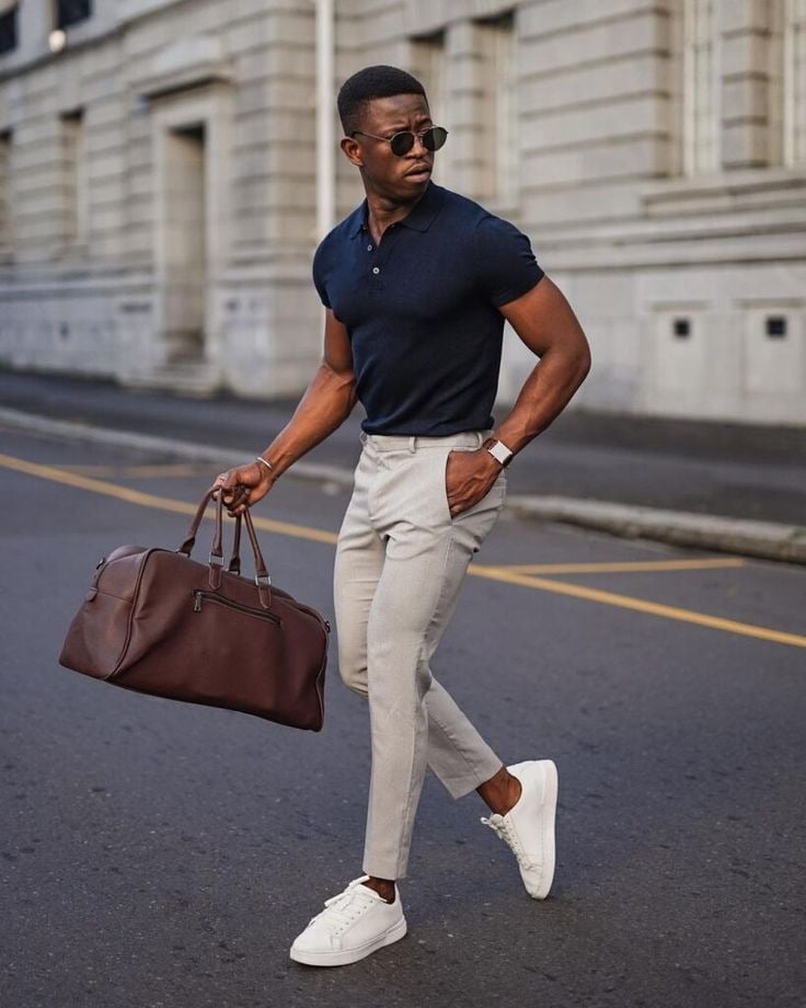 Stylish man in a navy polo and beige pants walking down the street with a brown leather bag, wearing sunglasses and white