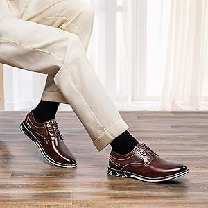 Man wearing stylish brown leather oxford shoes with beige trousers, sitting on wooden floor in bright modern room.