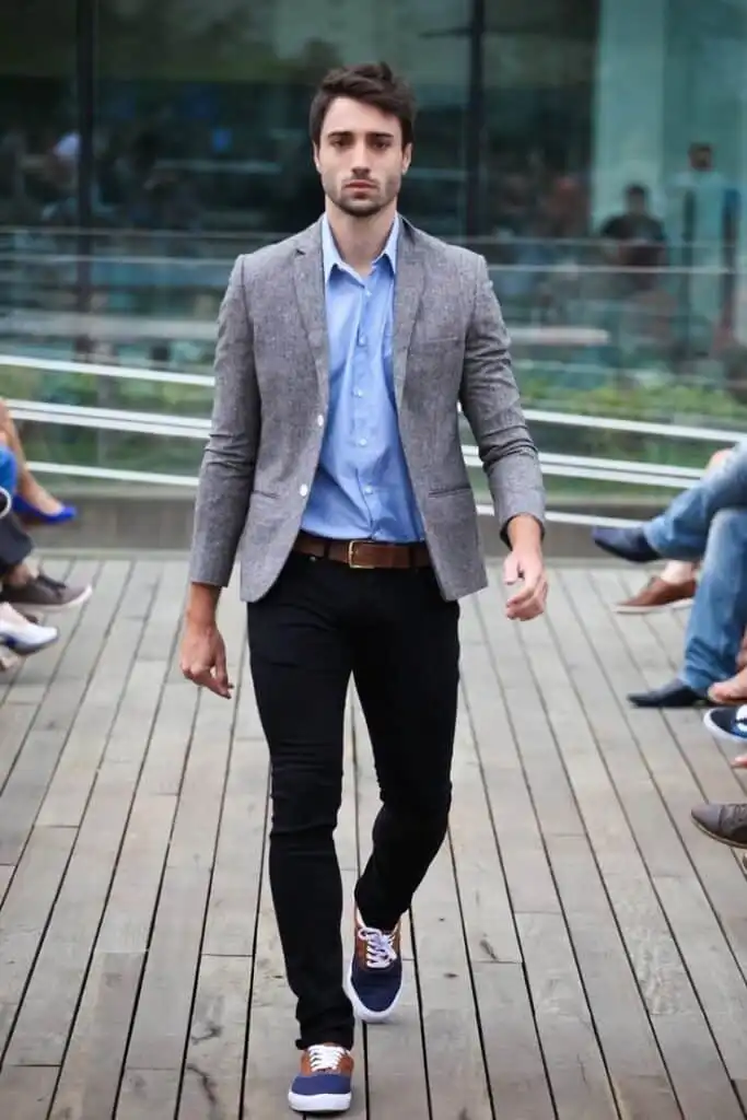 Man in stylish casual attire walking on a wooden deck, wearing a gray blazer, blue shirt, black pants, and sneakers.