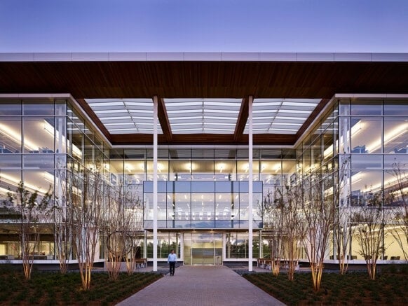 Modern glass building exterior at dusk with person walking towards entrance, featuring trees and a well-lit interior.