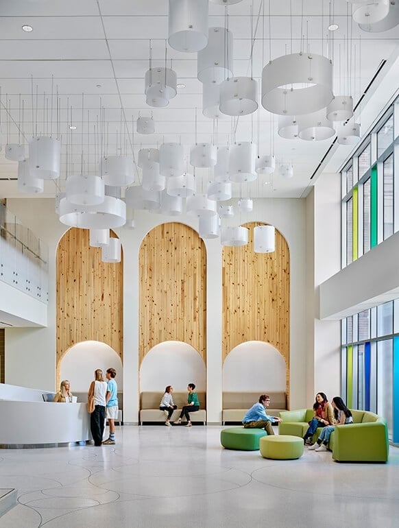 Modern lobby interior with people sitting. Features wooden arches, hanging lights, and colorful seating. Bright, spacious,