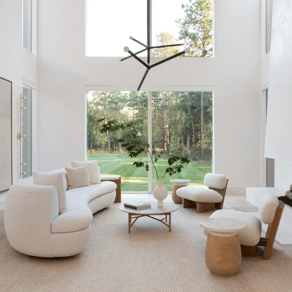Modern living room with elegant white furniture, large windows, and minimalist decor, overlooking a lush garden.