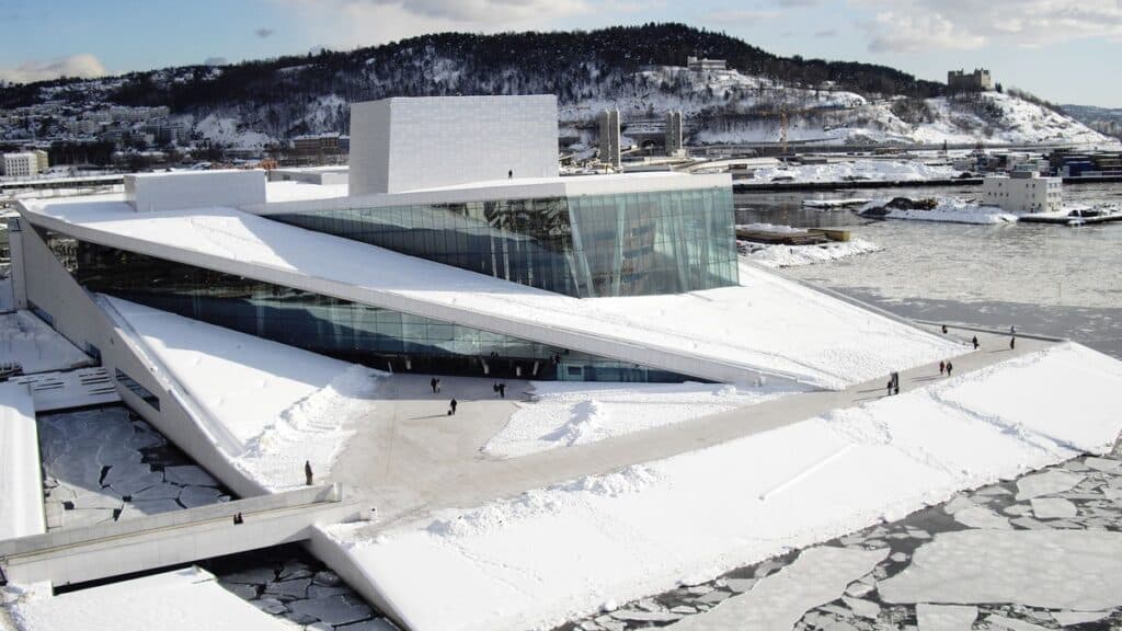 Snow-covered Oslo Opera House in Norway, featuring modern architecture by the waterfront, with people walking in the winter