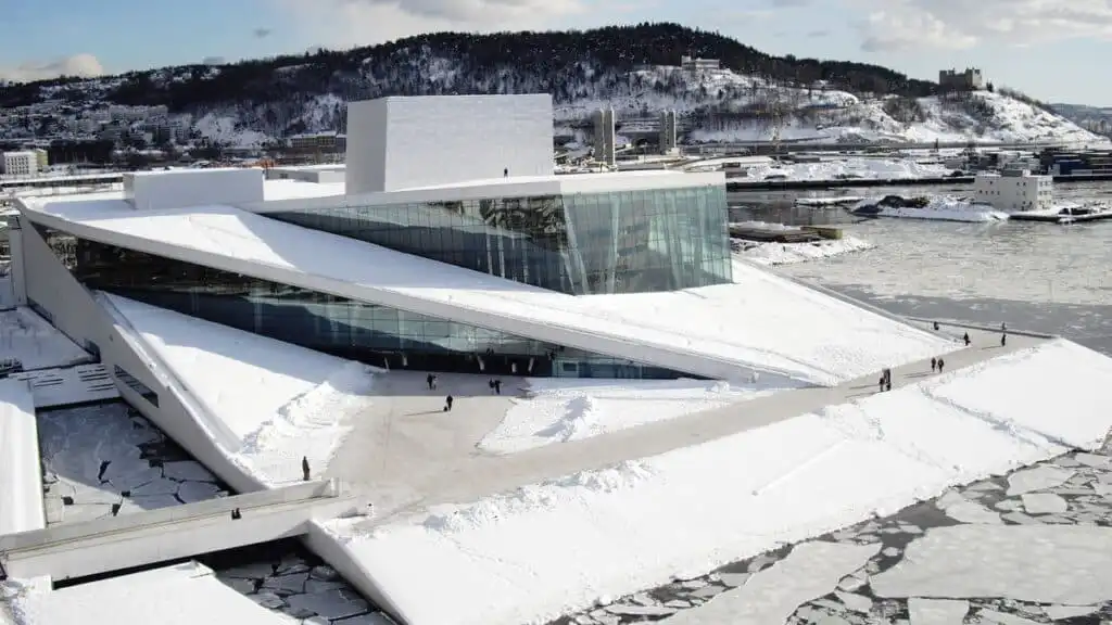 Snow-covered Oslo Opera House in Norway, featuring modern architecture by the waterfront, with people walking in the winter