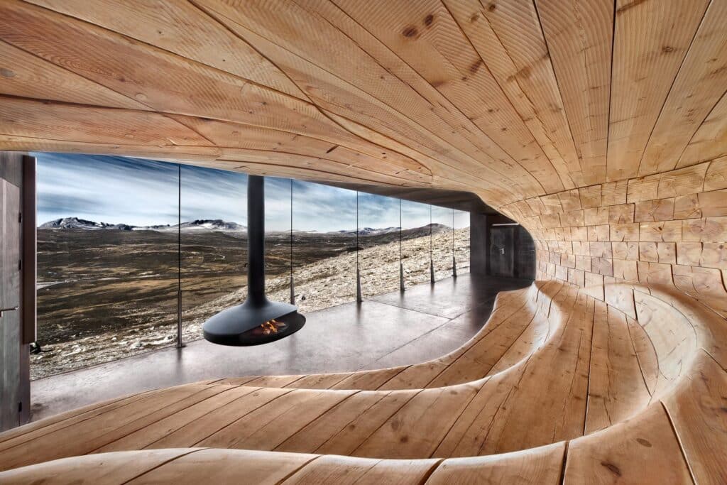 Modern wooden interior with sweeping curves and a suspended fireplace, overlooking a vast mountain landscape through