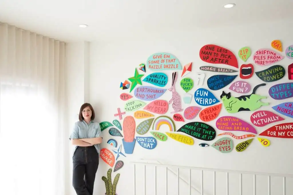 Woman standing by a colorful wall art full of quirky, playful text and shapes in a bright room, creating a lively and