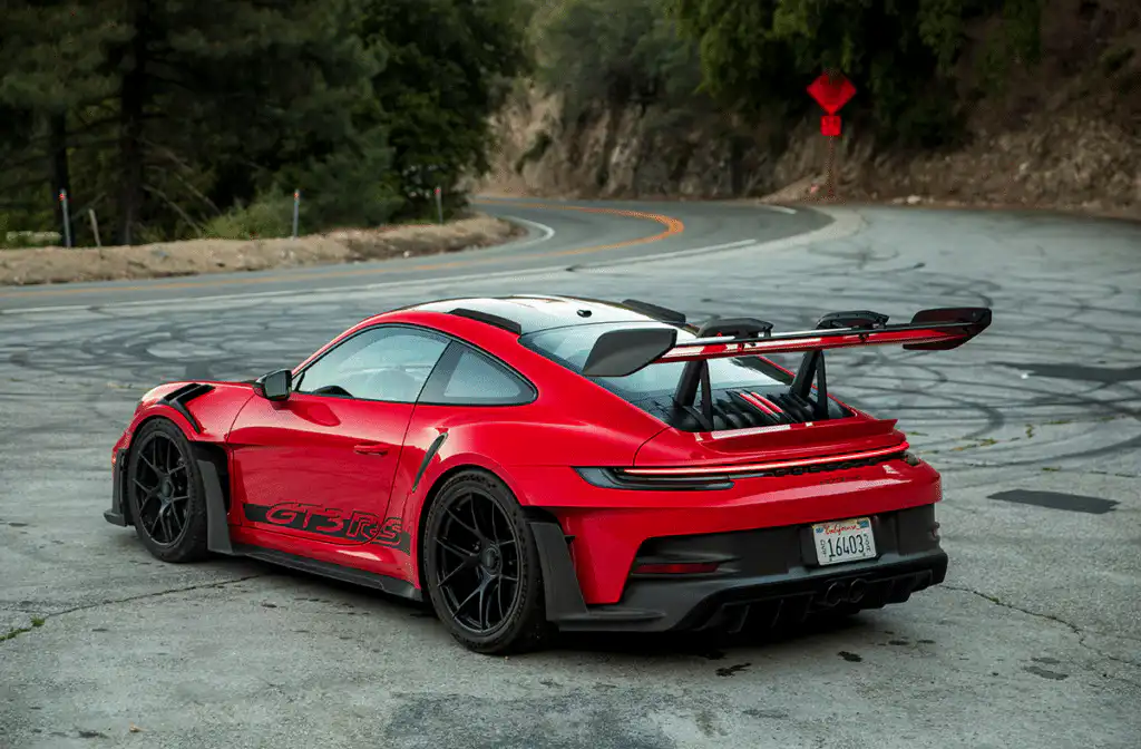 Red sports car, parked at a curvy mountain road with trees, showcasing sleek design and performance features from rear angle.