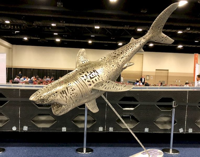 Intricate metal shark sculpture displayed at an indoor event, showing detailed cut-out designs, with people gathered in the
