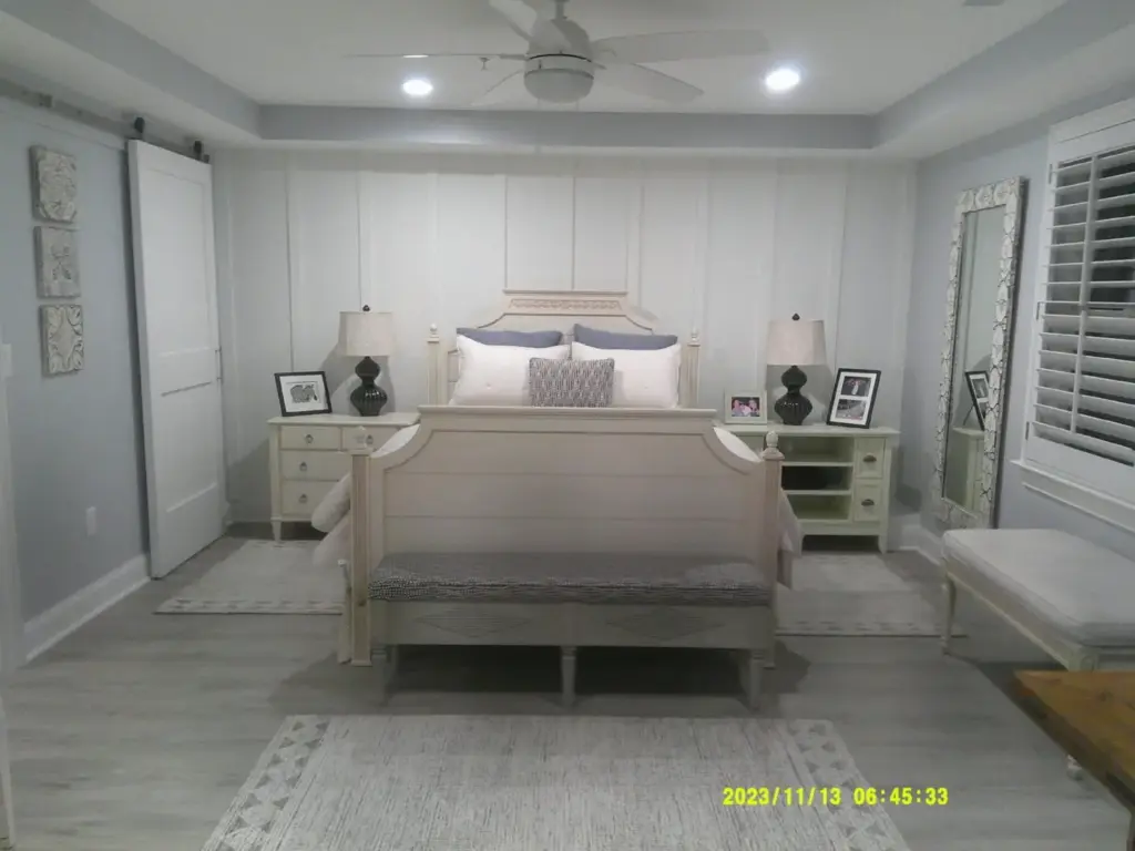 Modern bedroom with white bed, decorative lamps, and framed photos, featuring light grey walls and a large mirror near the window.