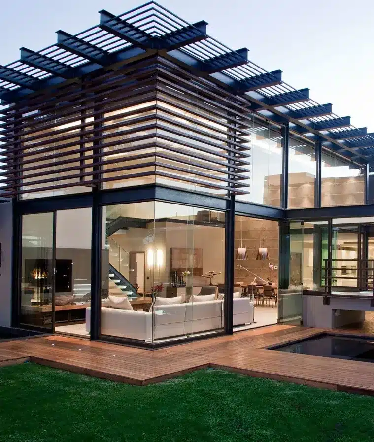 Modern glass house exterior with wooden accents, lush green lawn, and cozy interior view, showcasing contemporary