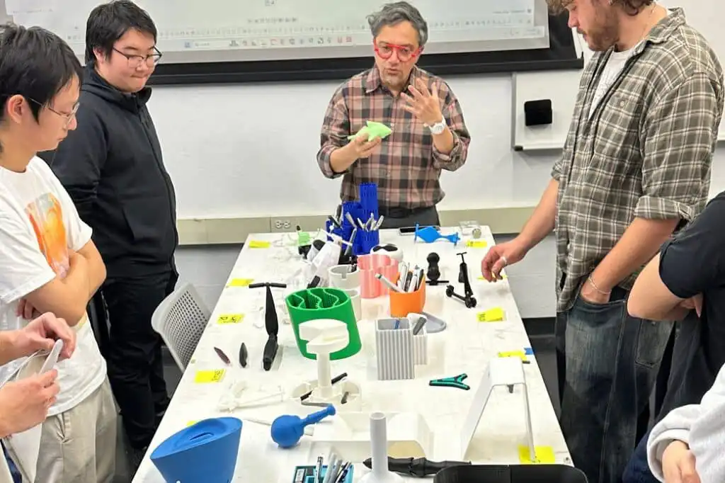 Man demonstrating 3D-printed designs to a group at a workshop table, highlighting technology and innovation.