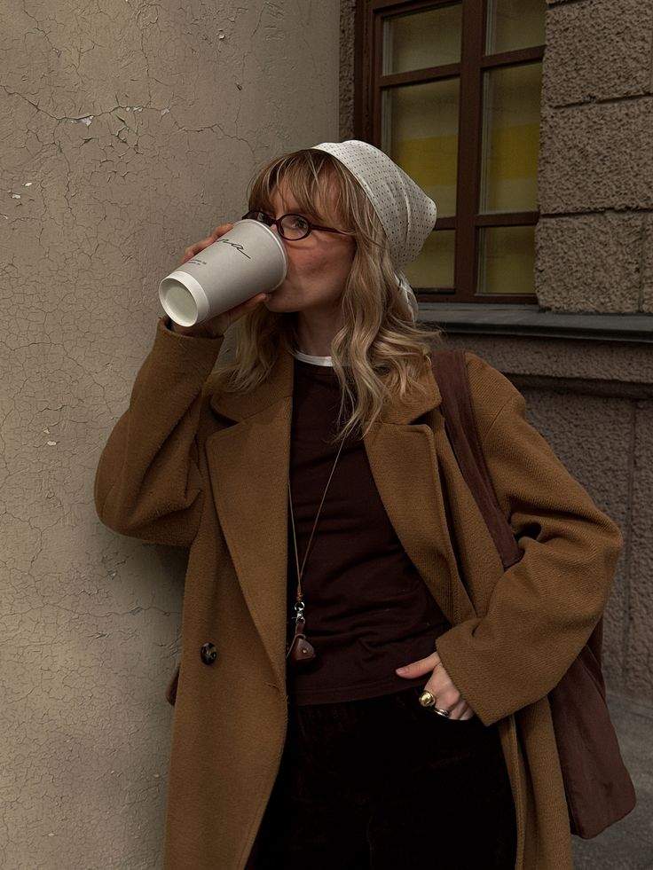 Woman in a brown coat and white headscarf drinks coffee against a textured wall. Casual city street style, fall fashion.
