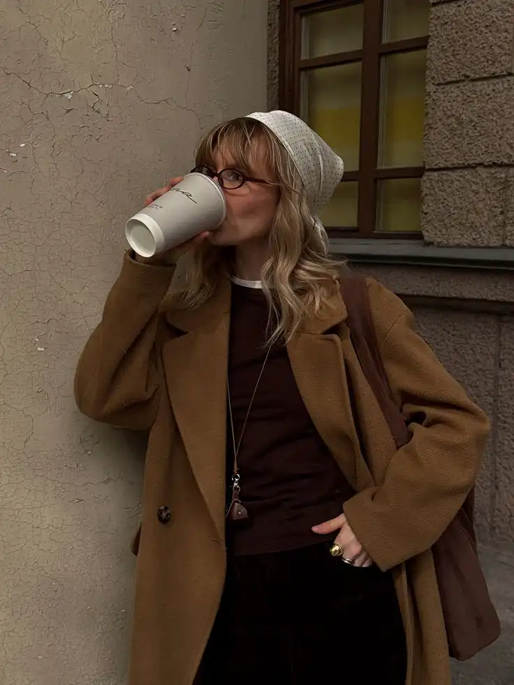 Woman in a brown coat and white headscarf drinks coffee against a textured wall. Casual city street style, fall fashion.