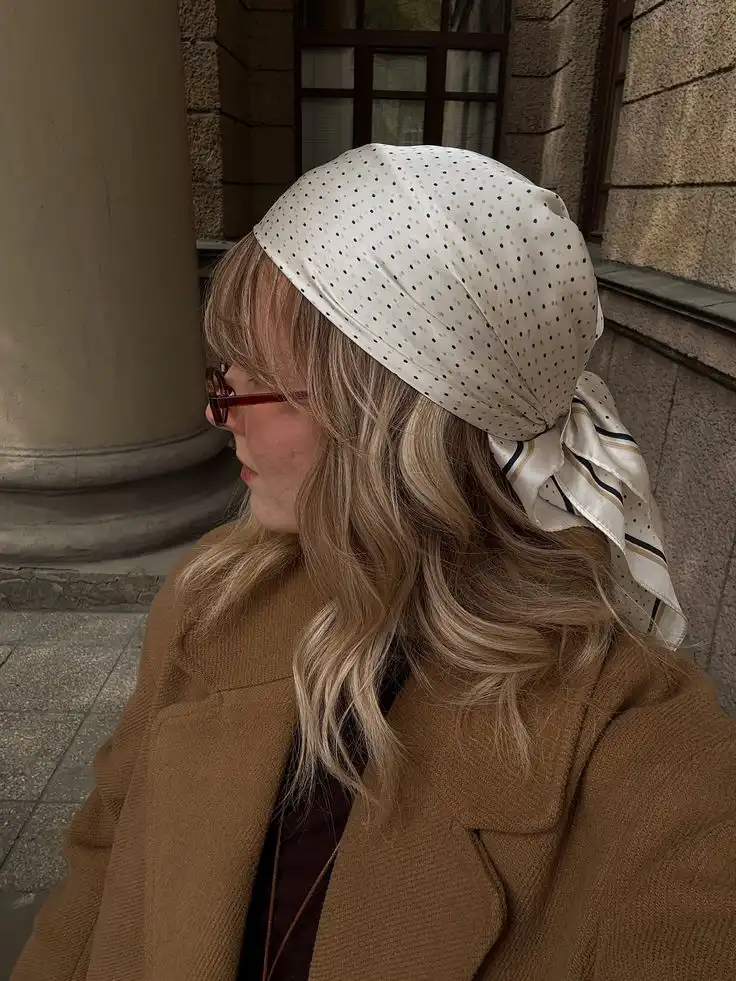 Woman in a brown coat and polka dot headscarf looks sideways, standing near stone building.