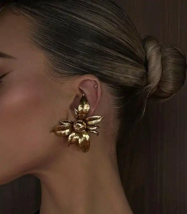 Woman with sleek bun wearing large gold flower-shaped earring, side profile with wooden background. Fashionable and elegant jewelry design.