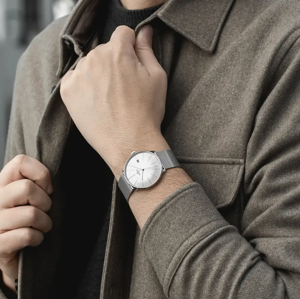 Man wearing a sleek silver wristwatch with a mesh band and a brown coat, adjusting his collar stylishly.
