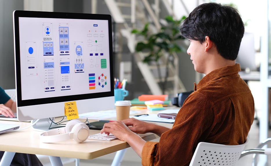 Designer working on a computer with UI/UX software, developing app prototypes in a modern office setting.