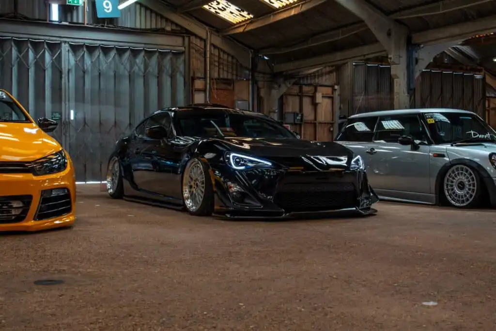 Sleek black sports car with custom rims showcased indoors alongside yellow and silver vehicles in a warehouse setting.