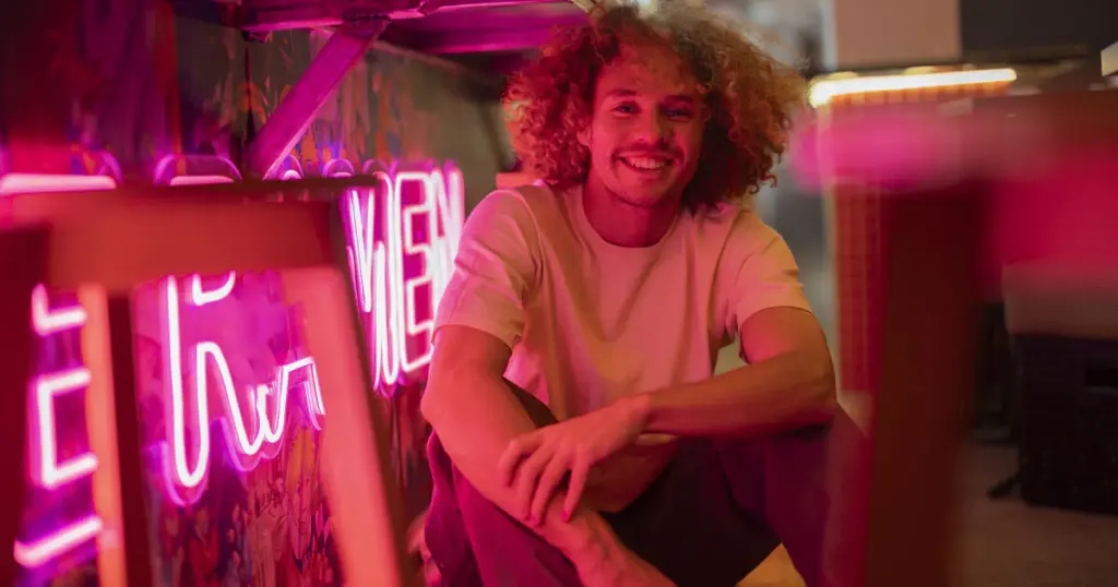 neon-sign-affects-the-mood-and-ambiance Smiling person with curly hair in neon-lit room, wearing a casual shirt, surrounded by vibrant pink lighting and neon signs. | Sky Rye Design Smiling person with curly hair in neon-lit room, wearing a casual shirt, surrounded by vibrant pink lighting and neon signs.