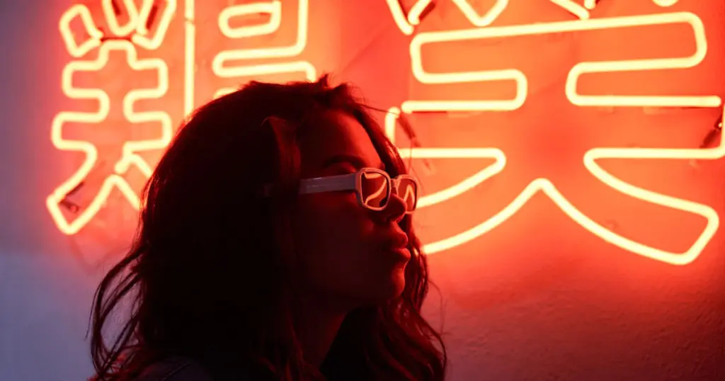 neon-sign-market-trends Woman in sunglasses stands before vibrant red neon sign with bold Asian characters, reflecting urban nightlife ambiance. | Sky Rye Design Woman in sunglasses stands before vibrant red neon sign with bold Asian characters, reflecting urban nightlife ambiance.