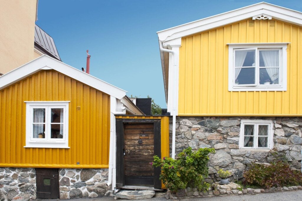 Two charming yellow wooden houses with stone foundations against a clear blue sky, featuring white framed windows and vivid greenery.