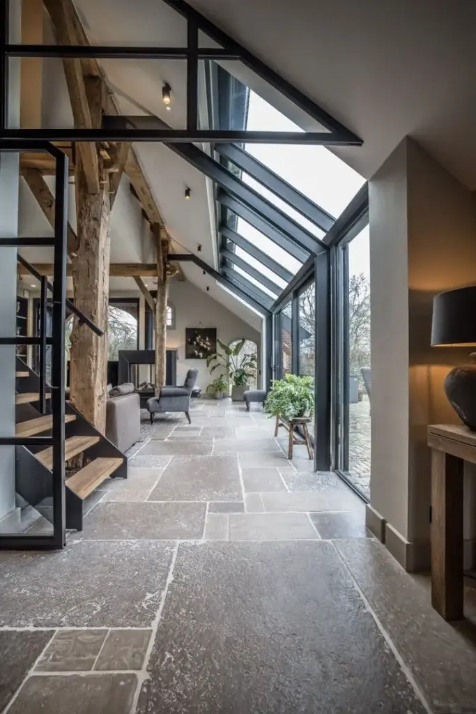 Modern rustic interior with large windows, stone floor, wooden beams, and cozy seating, blending nature with contemporary design.