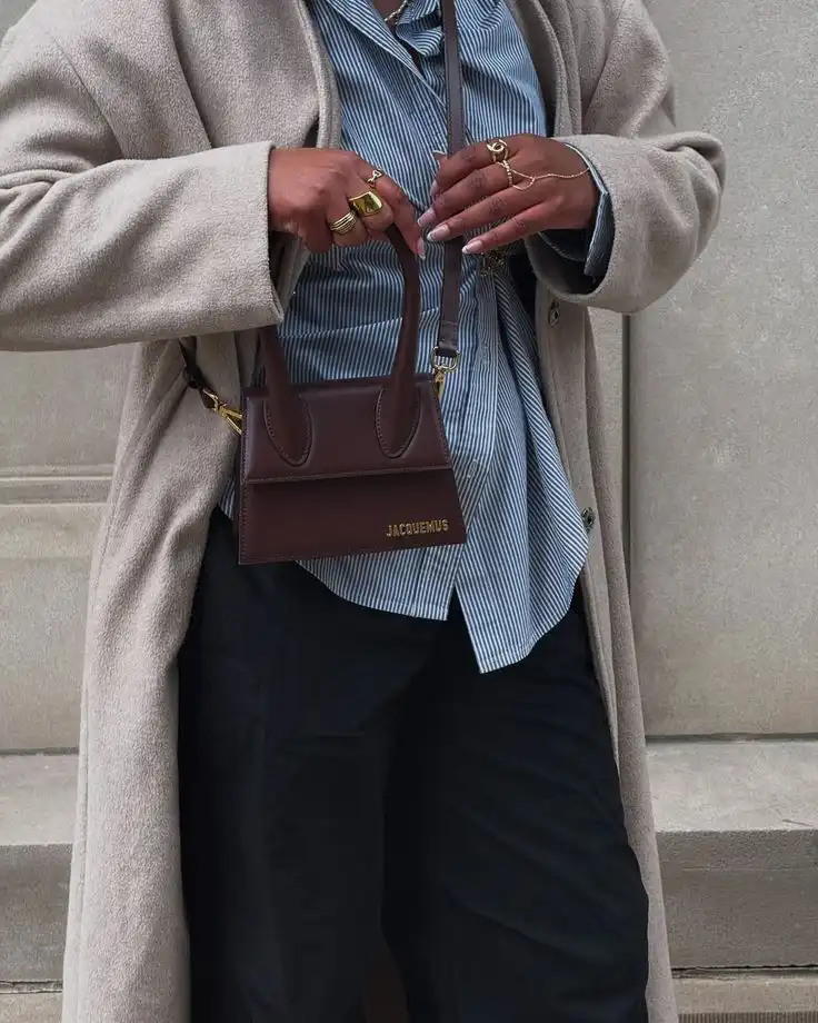 Stylish brown Jacquemus mini bag paired with a cozy beige coat and striped shirt, accessorized with gold rings.