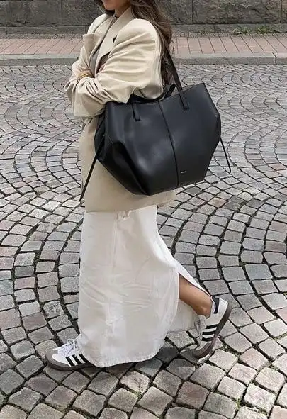 Woman in a beige coat with a black handbag and white skirt, walking on a cobblestone street, wearing Adidas sneakers. Fashionable street style.